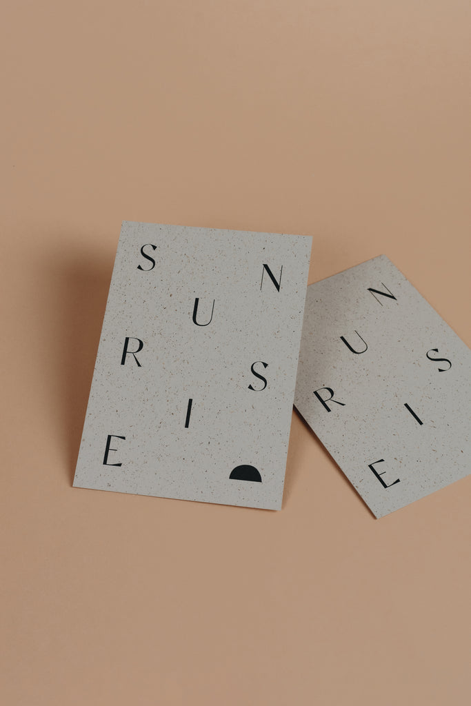 Sunrise postcard with black and light grey grass paper material and black sunrise text on it.